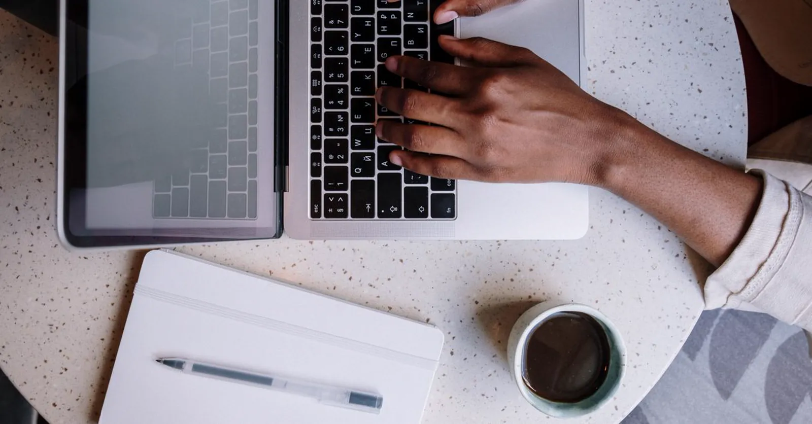 A small-business owner writing content on a laptop, a notebook and coffee beside them.