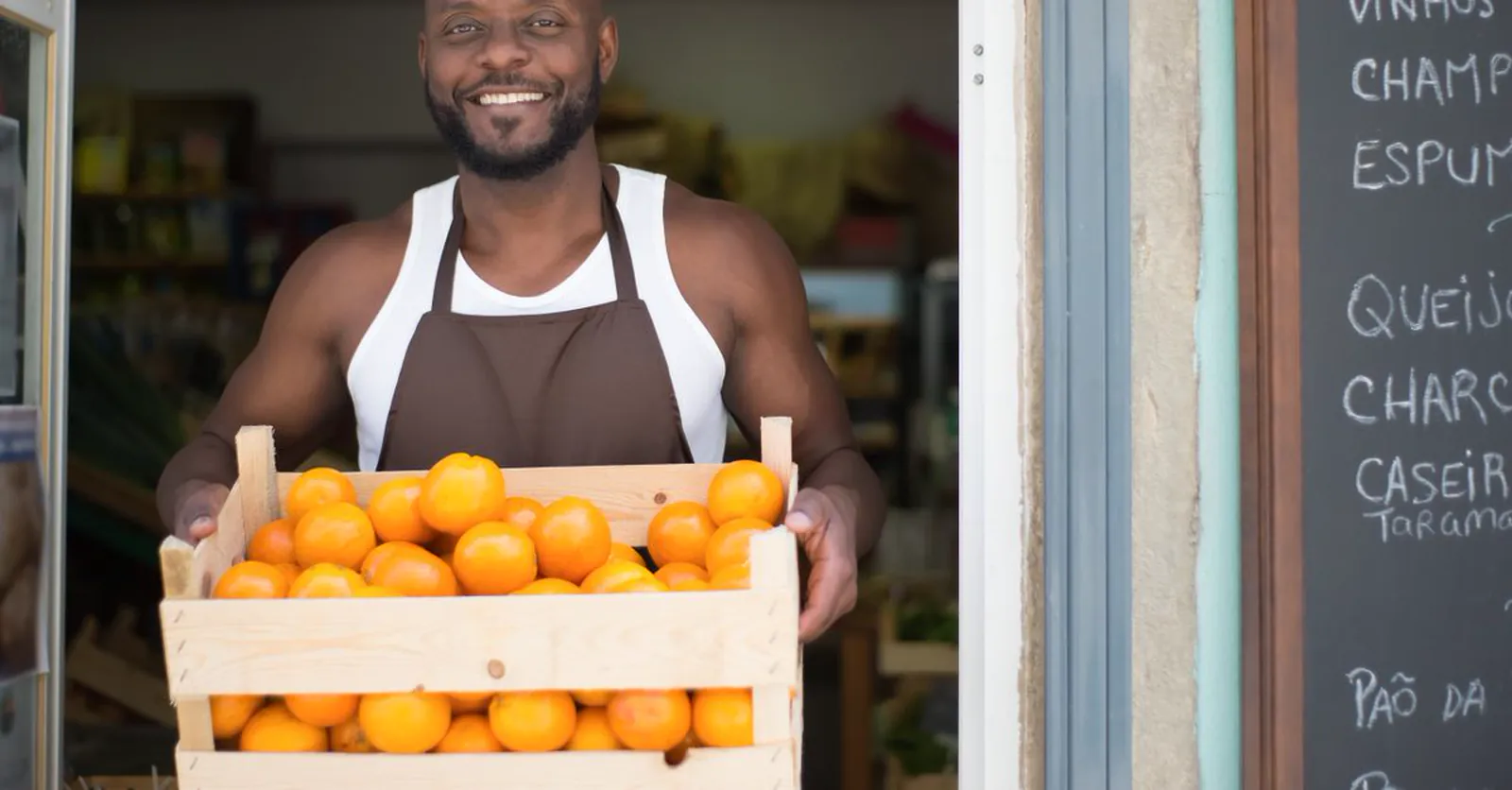A smiling shop owner in an apron at his storefront, holding a crate of fresh produce.