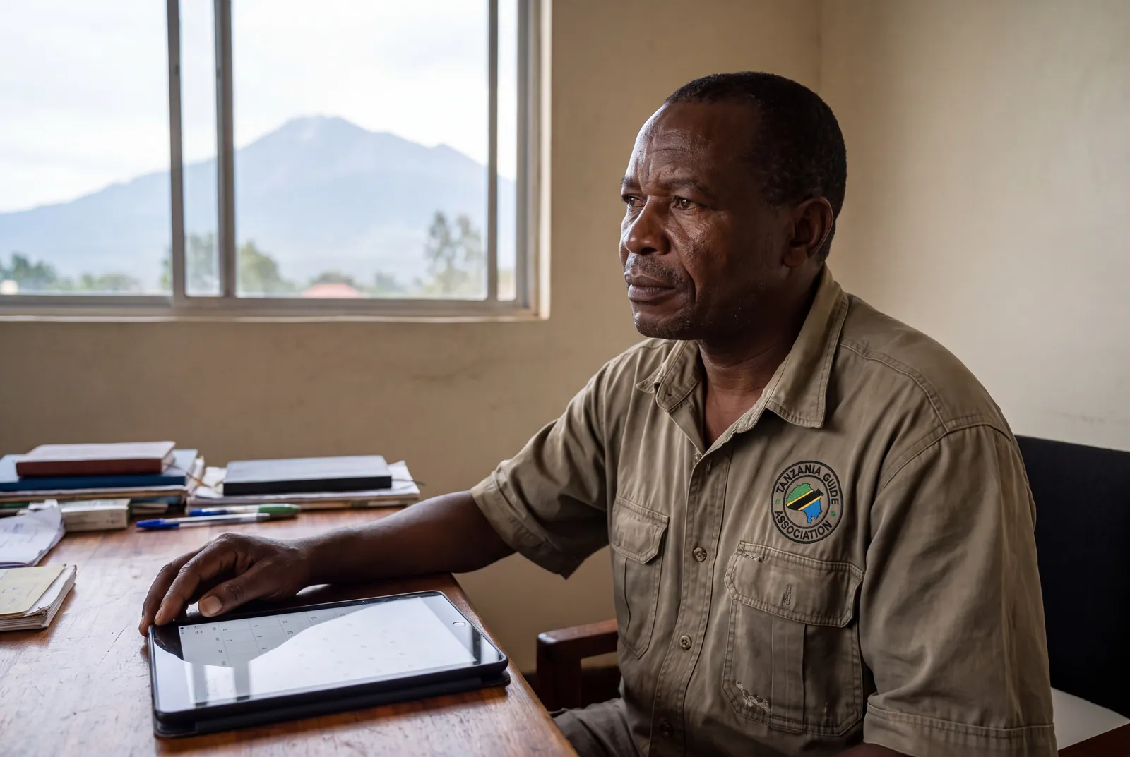 A Tanzanian safari operator checking a booking calendar on a tablet, Mount Meru framed in the window behind him.